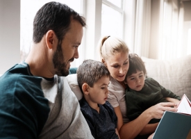 Family with book, reading with parents and children, happiness at home, story time and learning. Love, relationship and happy people bonding and care in living room with education and development Parents sitting on the couch with two young children reading them a book