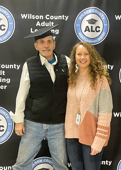Adult Education Man wearing a mortarboard stands next to a woman with a banner that says Wilson County Adult Learning Center in the background
