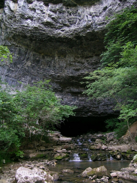 A large rock overhang with a stream running out of a cave entrance at its base